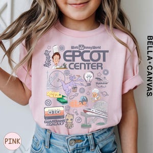 A young girl wearing a pink t-shirt with various Disney-themed graphics and characters, including a chef, a rocket ship, and other iconic symbols.