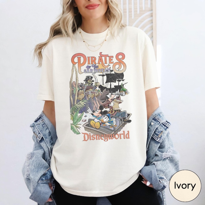 A woman wearing a white t-shirt with a graphic of Disney characters and text, standing in front of a white wall.