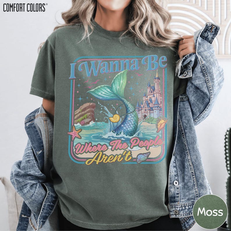 A person wearing a green t-shirt with a colorful graphic design featuring a mermaid, castle, and text.
