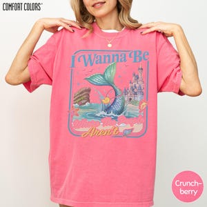 A woman wearing a pink t-shirt with a mermaid graphic and the text "I Wanna Be Arent" printed on it.