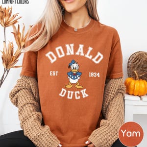 A woman wearing a brown t-shirt with a cartoon duck and text, along with a sweater and a pumpkin decoration, is smiling.