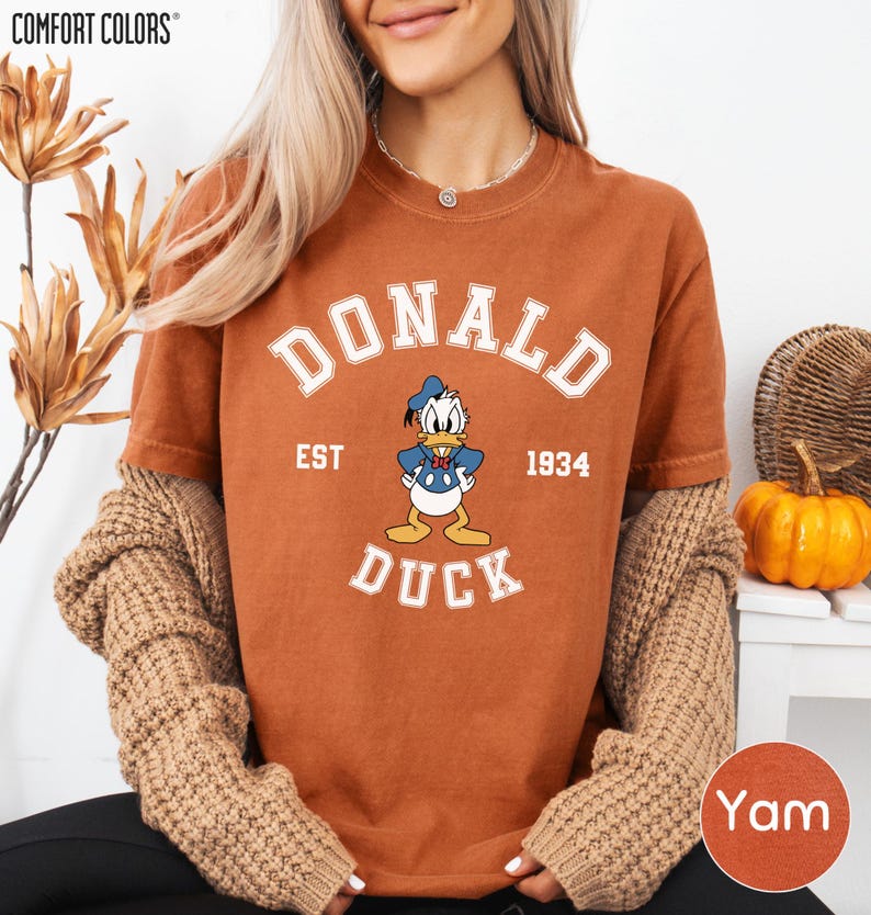 A woman wearing a brown t-shirt with a cartoon duck and text, along with a sweater and a pumpkin decoration, is smiling.