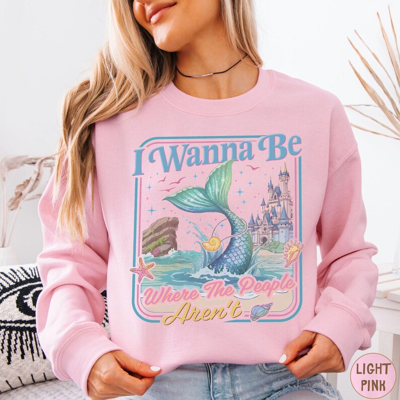 A woman wearing a pink sweatshirt with a graphic design featuring a mermaid, castle, and text.