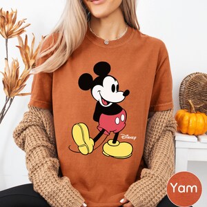 A woman wearing a brown t-shirt with a Mickey Mouse graphic and yellow shoes, sitting in front of a plant and a small pumpkin.