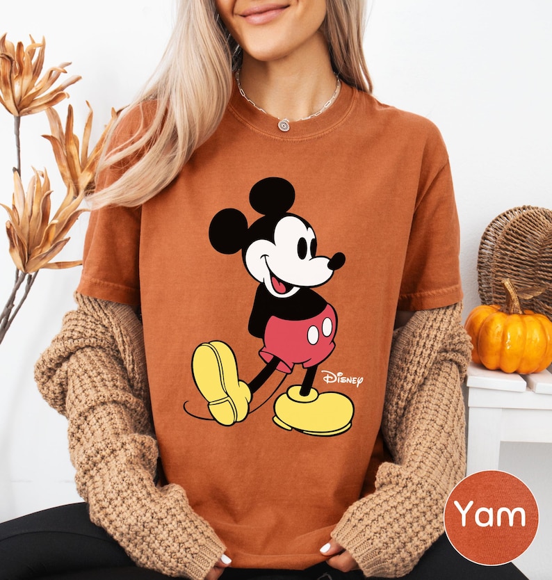 A woman wearing a brown t-shirt with a Mickey Mouse graphic and yellow shoes, sitting in front of a plant and a small pumpkin.