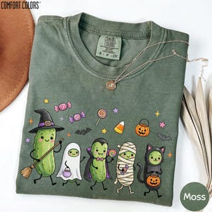 a green t-shirt with a Halloween-themed design, including a witch, a mummy, a pumpkin, and other spooky and festive characters.