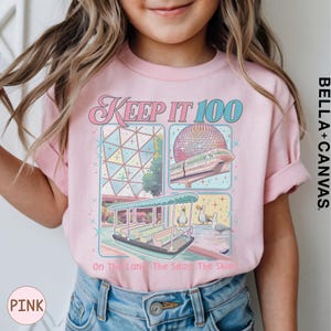 A young girl wearing a pink t-shirt with a graphic design featuring a Ferris wheel and a boat, along with the text "Keep it 100 On the land The sea The skies" printed on it.