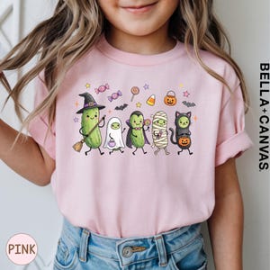 A young girl wearing a pink t-shirt with a Halloween-themed graphic of various cartoon characters, including a witch, pumpkin, ghost, mummy, and cat.
