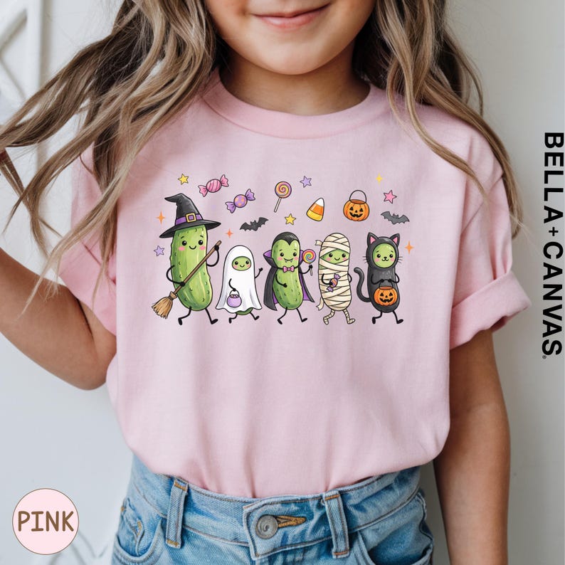 A young girl wearing a pink t-shirt with a Halloween-themed graphic of various cartoon characters, including a witch, pumpkin, ghost, mummy, and cat.
