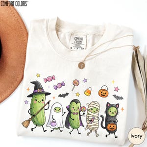A white t-shirt with a Halloween-themed graphic design featuring a group of anthropomorphic vegetables, including a witch, pumpkin, cat, and ghost, along with various Halloween-related elements such as bats, candy, and a pumpkin.