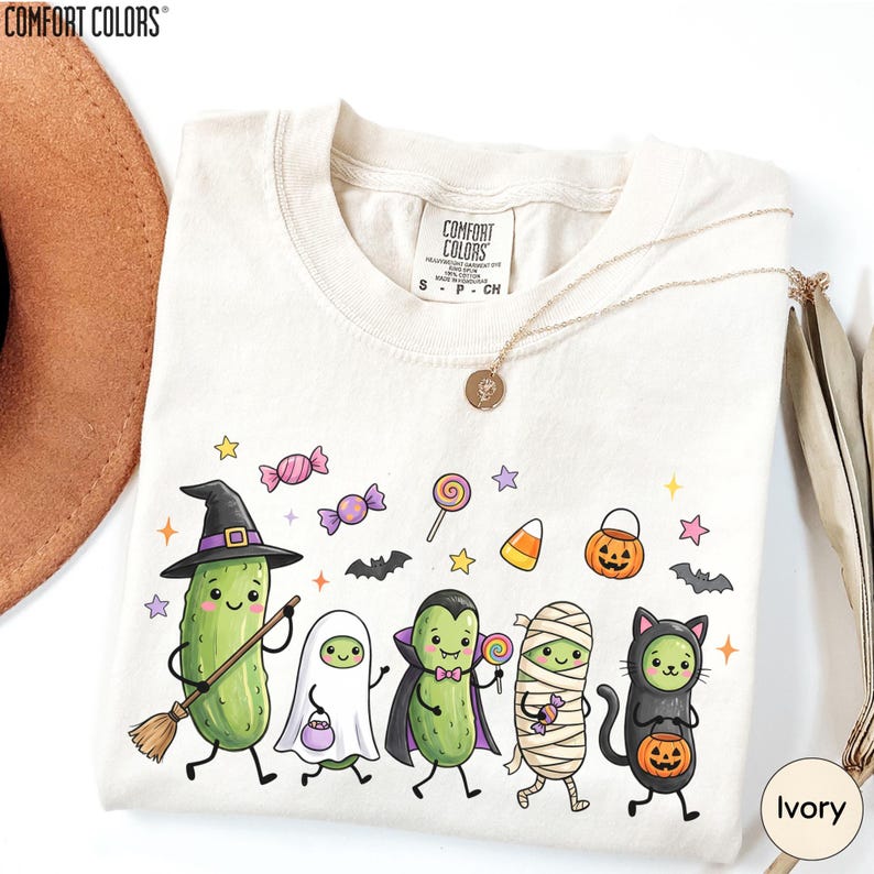 A white t-shirt with a Halloween-themed graphic design featuring a group of anthropomorphic vegetables, including a witch, pumpkin, cat, and ghost, along with various Halloween-related elements such as bats, candy, and a pumpkin.