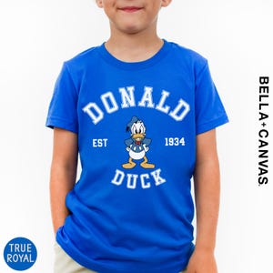 A young boy wearing a blue t-shirt with a cartoon image of Donald Duck and text that says "DONALD EST. 1934 DUCK".