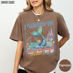 A woman wearing a brown t-shirt with a colorful graphic design featuring a mermaid, a castle, and the text "I Wanna Be Where The People Aren't".