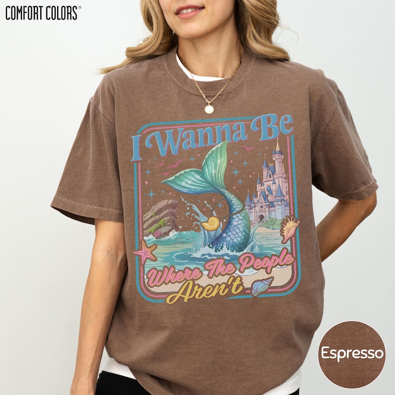 A woman wearing a brown t-shirt with a colorful graphic design featuring a mermaid, a castle, and the text "I Wanna Be Where The People Aren't".