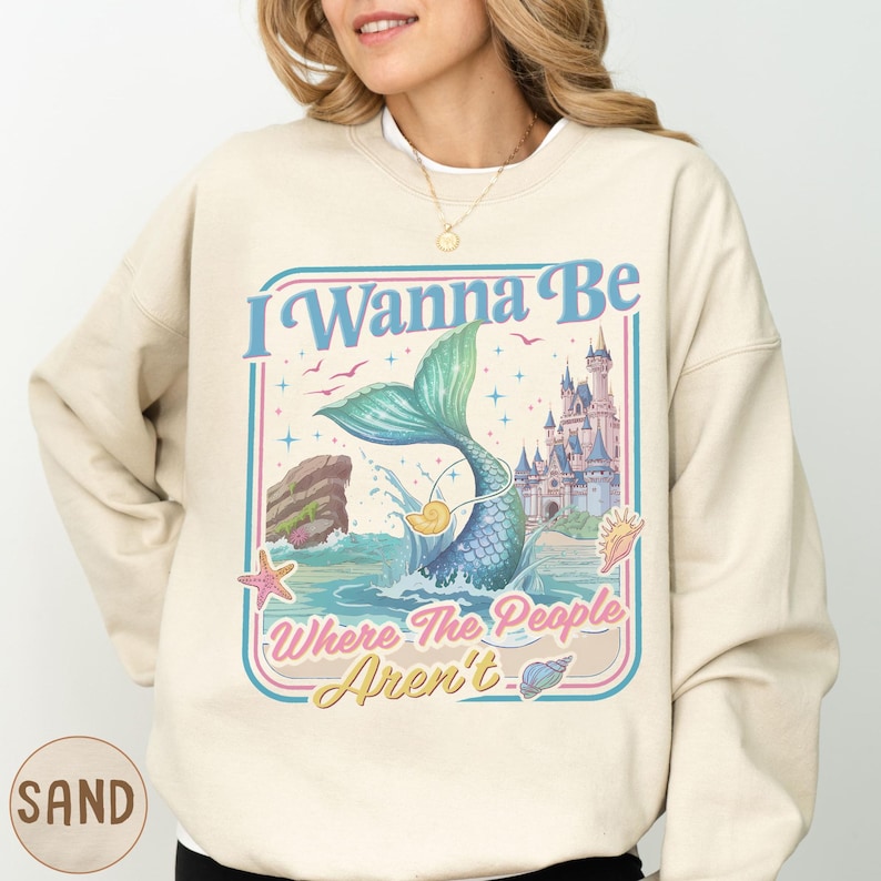 A woman wearing a beige sweatshirt with a colorful graphic design featuring a mermaid, a castle, and the text "I Wanna Be Where The People Aren't".