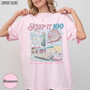 A woman wearing a pink t-shirt with a graphic design depicting a carnival or amusement park ride, along with text that reads "Keep It 100 On The Land. The Seas. The Skies".