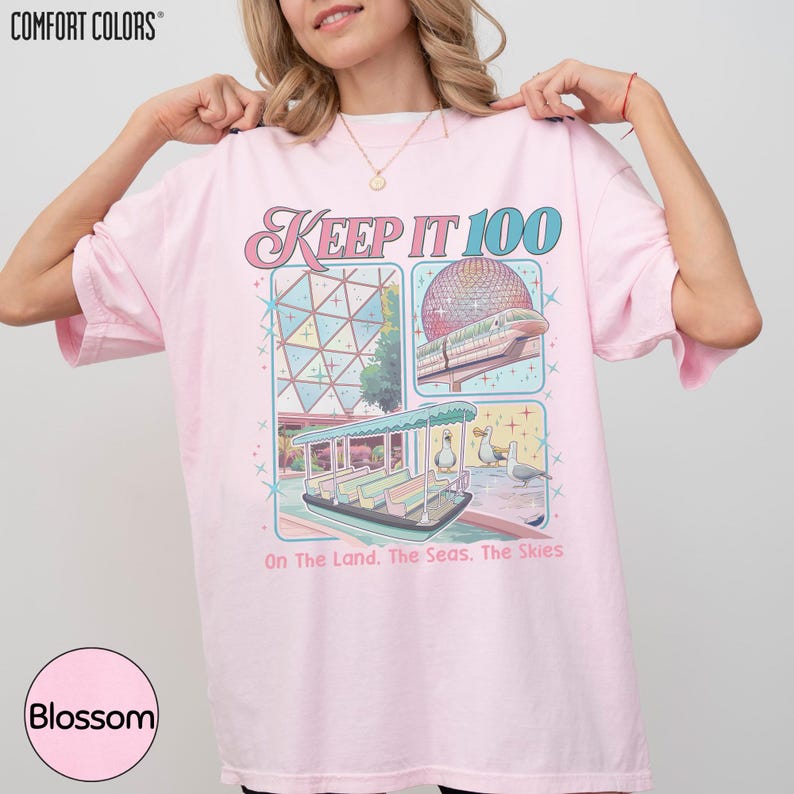 A woman wearing a pink t-shirt with a graphic design depicting a carnival or amusement park ride, along with text that reads "Keep It 100 On The Land. The Seas. The Skies".