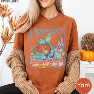 A woman wearing a brown t-shirt with a mermaid graphic and text, along with a sweater and a pumpkin decoration.