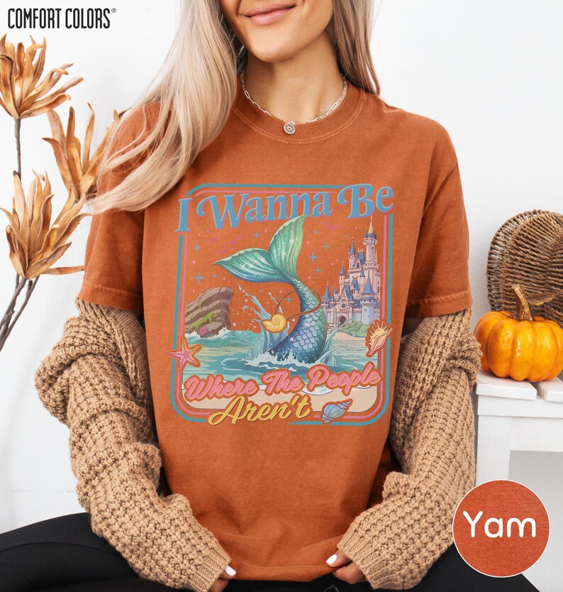 A woman wearing a brown t-shirt with a mermaid graphic and text, along with a sweater and a pumpkin decoration.