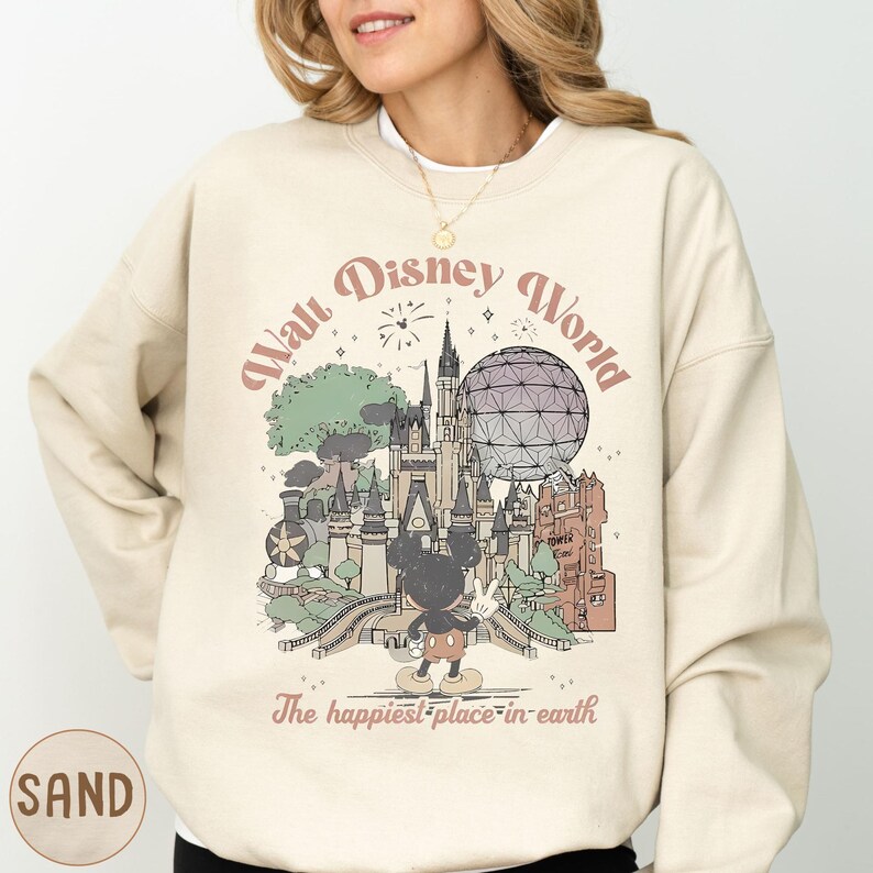 A woman wearing a beige sweatshirt with a graphic design depicting various Disney landmarks and a Mickey Mouse.
