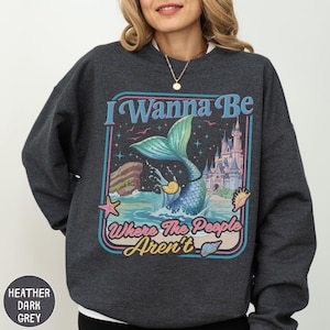 A woman wearing a gray sweatshirt with a colorful graphic design featuring a mermaid, a castle, and the text "I Wanna Be Where The People Aren't".