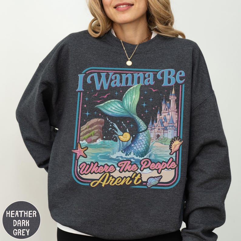 A woman wearing a gray sweatshirt with a colorful graphic design featuring a mermaid, a castle, and the text "I Wanna Be Where The People Aren't".
