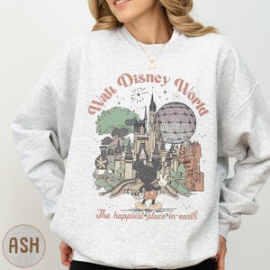 A woman wearing a gray sweatshirt with a graphic design depicting various Disney landmarks and characters, including Mickey Mouse.