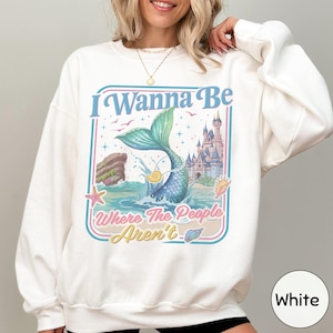 A woman wearing a white sweatshirt with a colorful graphic design featuring a mermaid, castle, and text.