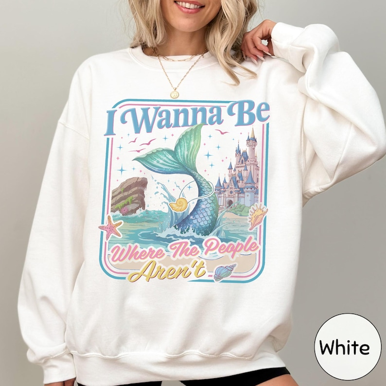 A woman wearing a white sweatshirt with a colorful graphic design featuring a mermaid, castle, and text.