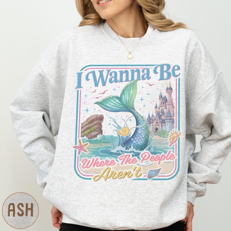 A woman wearing a gray sweatshirt with a colorful graphic design featuring a mermaid, castle, and text.