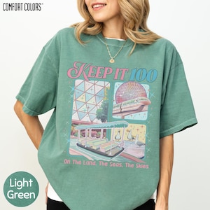 A woman wearing a green t-shirt with a graphic design featuring a large dome, a boat, and a beach scene.