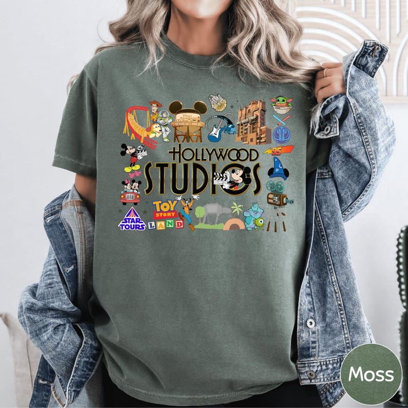 A person wearing a green t-shirt with various Disney and Pixar characters and logos, including Mickey Mouse, Minnie Mouse, Toy Story, Star Wars, and others, along with the text "Hollywood STUDIOS" and "Moss".