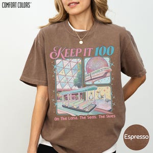 A woman wearing a brown t-shirt with a graphic design featuring a trolley car and a globe, along with the text "Keep It 100 On The Land. The Seas. The Skies" and "Espresso".
