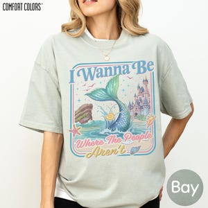 A woman wearing a gray t-shirt with a colorful graphic design featuring a mermaid, a castle, and the text "I Wanna Be Where The People Aren't".