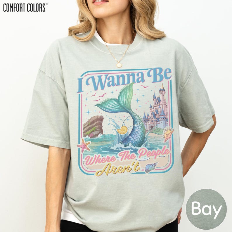 A woman wearing a gray t-shirt with a colorful graphic design featuring a mermaid, a castle, and the text "I Wanna Be Where The People Aren't".