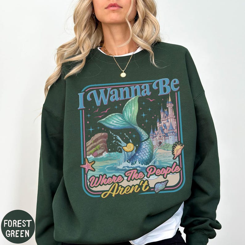 A woman wearing a green sweatshirt with a colorful graphic design featuring a mermaid, castle, and text.