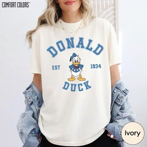 A woman wearing a white t-shirt with a graphic of a cartoon duck and text that says "DONALD DUCK EST 1934". She is also wearing a denim jacket and has her hands in her pockets.