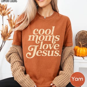 A woman wearing a brown t-shirt with the text "cool moms love Jesus" and beige cardigan sleeves, sitting in front of a plant and a pumpkin decoration.