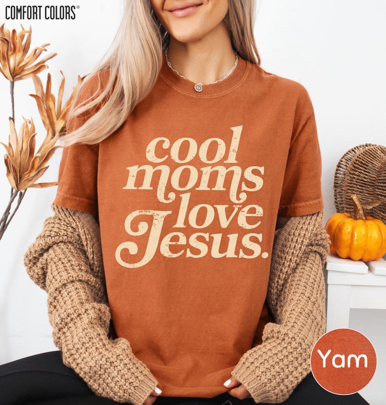 A woman wearing a brown t-shirt with the text "cool moms love Jesus" and beige cardigan sleeves, sitting in front of a plant and a pumpkin decoration.