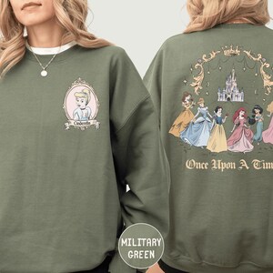 A woman wearing a green sweatshirt with a graphic of a group of princesses and a castle on the back.