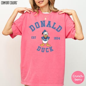 A woman wearing a pink t-shirt with a graphic of a cartoon duck and text that says "DONALD DUCK EST 1934".