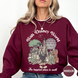 A woman wearing a maroon sweatshirt with a graphic design depicting various Disney landmarks and the text "The happiest place in earth".