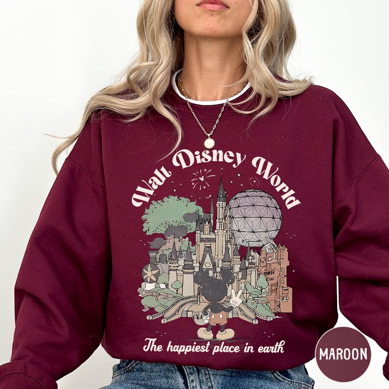 A woman wearing a maroon sweatshirt with a graphic design depicting various Disney landmarks and the text "The happiest place in earth".