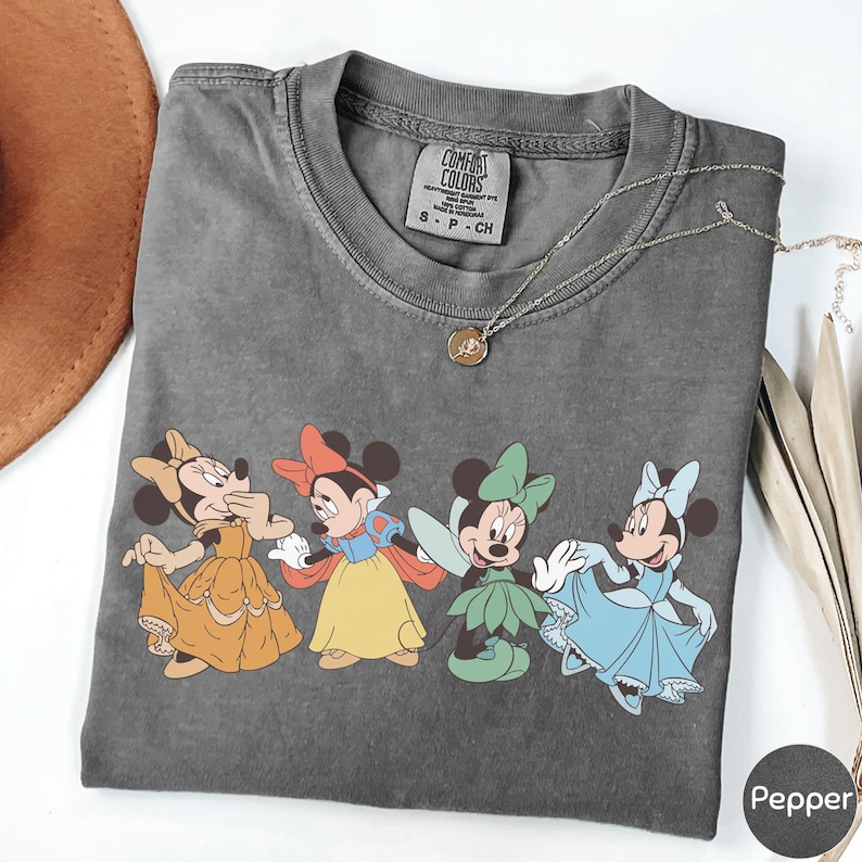 A gray t-shirt with a graphic of four cartoon Minnie Mouse characters in different outfits, including a yellow dress, a blue dress, a green dress, and a pink dress.