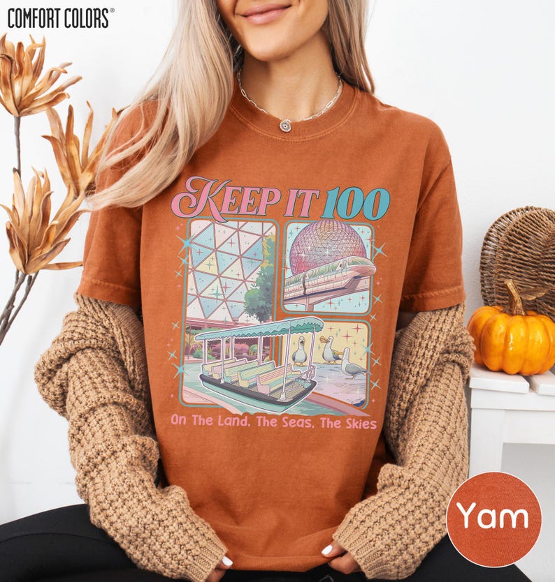 A woman wearing a t-shirt with a graphic design of a trolley car and the text "Keep it 100 On the land, the seas, the skies" is sitting in front of a plant and a pumpkin.