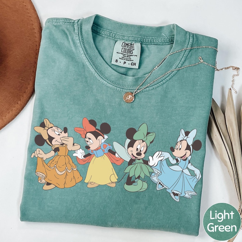 A green t-shirt with a printed design of Minnie Mouse and Snow White characters, along with a necklace and a brown hat.