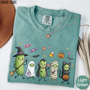 a green t-shirt with a Halloween-themed design, including a witch, a mummy, a pumpkin, a cat, and a cucumber.