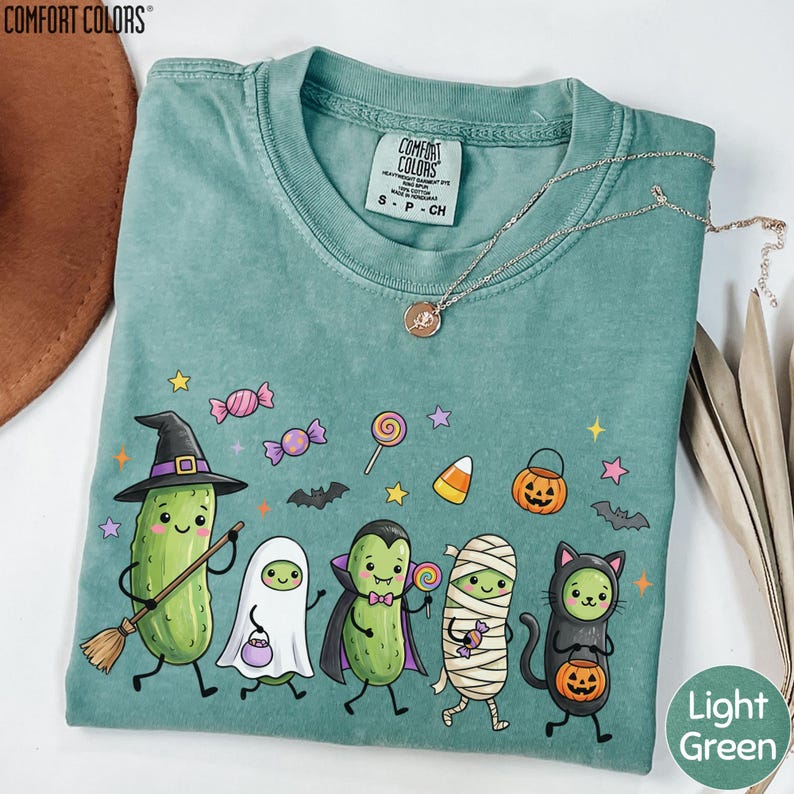a green t-shirt with a Halloween-themed design, including a witch, a mummy, a pumpkin, a cat, and a cucumber.