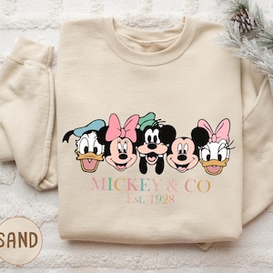 May include: A sand-colored sweatshirt featuring a graphic of Disney characters: Donald Duck, Minnie Mouse, Goofy, Mickey Mouse, and Daisy Duck. The text "Mickey & Co. Est. 1928" is printed below the characters in pastel colors.