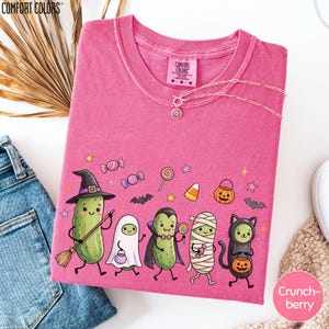 a pink t-shirt with a cartoon-style illustration of a group of anthropomorphic vegetables dressed in Halloween costumes, including a witch, pumpkin, skeleton, and ghost.